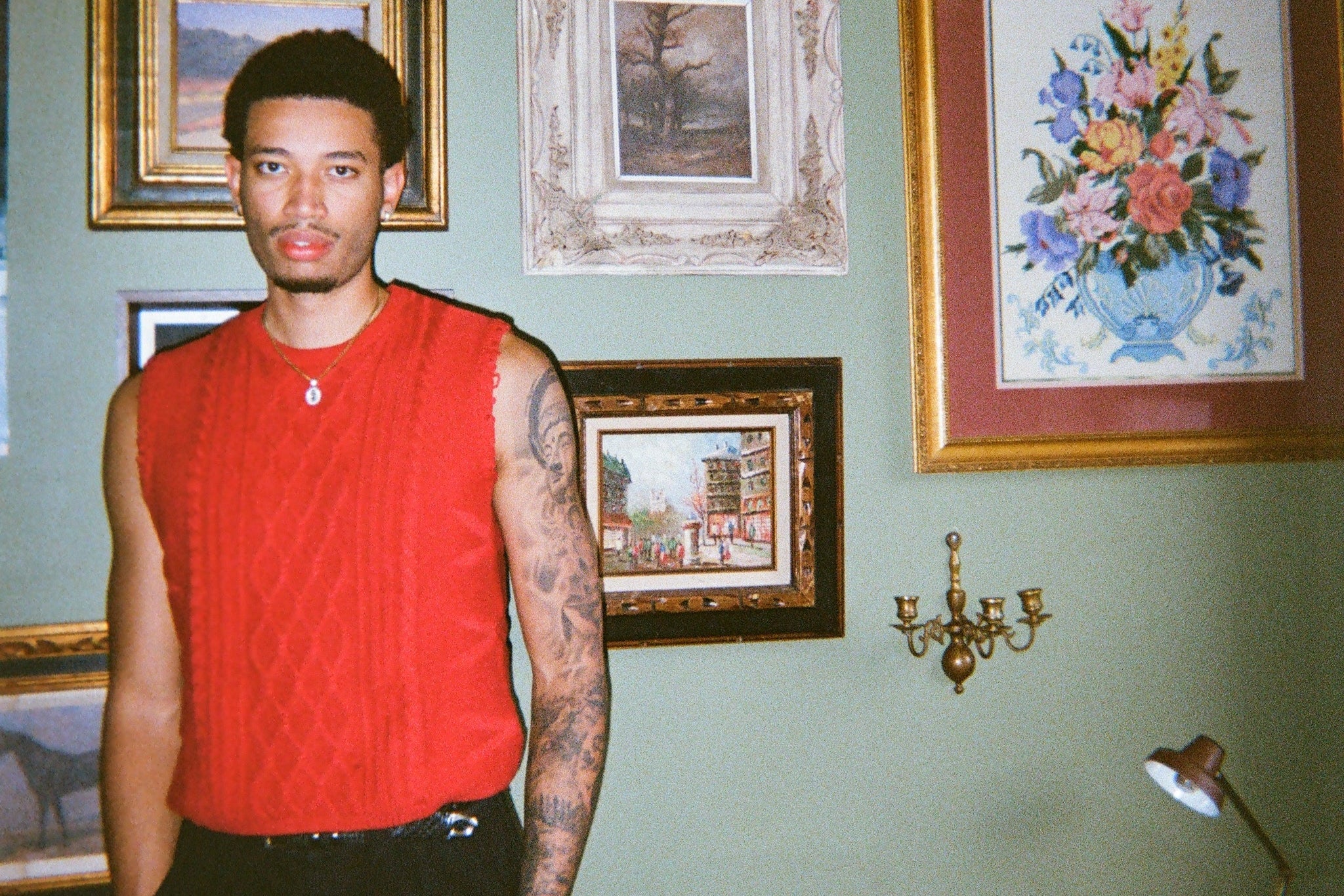 Person wearing a red knitted vest and black pants standing in a room with framed pictures on the wall.